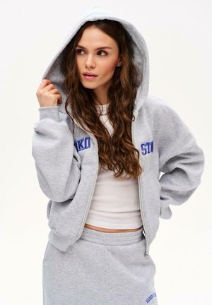 Młoda kobieta o długich brązowych włosach, ubrana w szarą bluzę z kapturem i pasującą spódnicę, trzymająca kapturem jedną ręką, patrząca w bok.