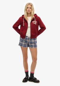 Sudadera con cremallera en color marrón y logo blanco, llevada sobre una camiseta blanca. Combinada con pantalones cortos de cuadros en azul y blanco. Zapatos negros y calcetines grises con rayas rojas.