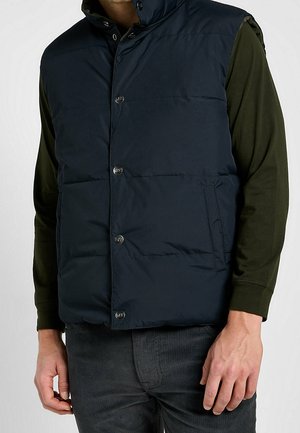 Mężczyzna ubrany w ciemnoniebieską pikowaną kamizelkę na oliwkowozielonej koszuli z długim rękawem i ciemnoszarych spodniach, stojący z rękami swobodnie opuszczonymi.