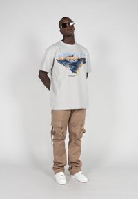 Lys grå grafisk t-skjorte med et ørnerdesign og blå/orange tekst, kombinert med beige cargo-bukser og hvite sneakers, som viser en avslappet stil.