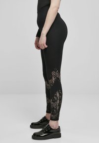 Leggings noirs avec des inserts en dentelle au niveau des mollets, présentant une coupe ajustée et une texture lisse, associés à des chaussures noires brillantes.
