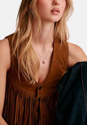 Junge Frau trägt eine braune, mit Fransen verzierte Wildlederweste und eine silberne Halskette mit einem roten Herzanhänger, hält dunklen Denimstoff in der Hand.