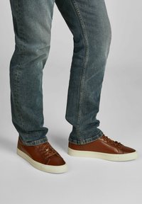 Mann, der blaue Jeans und braune Ledersneaker mit weißen Sohlen trägt, steht auf einem grauen Boden.
