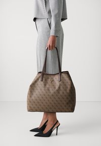 Bruine tas met patroon en donkerbruine handvatten, voorzien van een driehoekig logo. Gecombineerd met een zwart-wit geruit outfit en zwarte hakken.