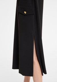 Schwarze Rock aus glattem Stoff mit einem seitlichen Schlitz und einer Tasche mit einem goldenen Knopfakzent. Das Design ist elegant und minimalistisch.