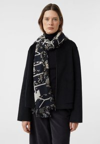 Kobieta w czarnej kurtce i spodniach, z wzorzystym czarno-białym szalem owiniętym wokół szyi, na jednolitym tle.