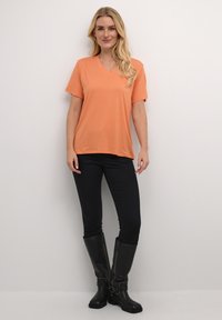 Orange v-neck t-shirt in lightweight fabric, short sleeves, paired with black fitted pants and knee-high black boots with buckle detail.