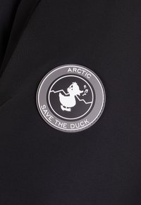 Okrągła łatka z materiału w kolorze czarnym, z białą ilustracją kaczki, konturem gór i napisem „ARCTIC SAVE THE DUCK” w układzie okrągłym.