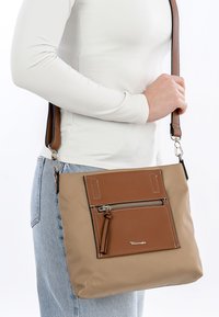 Tan axelväska med en framficka i brunt läder, zippered stängning, bårt strap med två färger och vit sömnadsdetalj. Slät textur.
