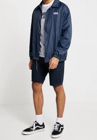 Rompevientos azul marino con cuello y botones, con un logo, combinado con una camisa a rayas, pantalones cortos azul marino y zapatillas negras.