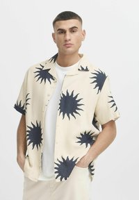 Camicia a maniche corte, con bottoni, in tessuto beige, caratterizzata da grandi motivi a raggi di sole scuri. Il colletto è aperto, rivelando una maglietta bianca sottostante.