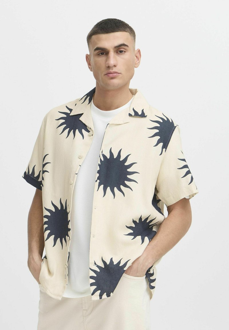 Camicia a maniche corte, con bottoni, in tessuto beige, caratterizzata da grandi motivi a raggi di sole scuri. Il colletto è aperto, rivelando una maglietta bianca sottostante.