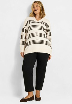 Mujer sonriente que lleva un jersey blanco con rayas negras, pantalones negros y bailarinas marrones, de pie frente a un fondo claro y liso.
