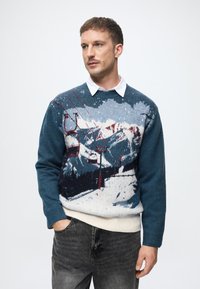 Zila džemperis ar kalnu ainavu un slēpošanas pacelšanas dizainu, ar baltu ribotu jostasvietu. Saskaņots ar tumši pelēkiem džinsiem.