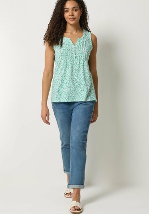 Vrouw met krullend haar, gekleed in een mouwloze groene blouse met patroon, opgerolde blauwe jeans en witte sandalen, die vooruit loopt tegen een effen achtergrond.