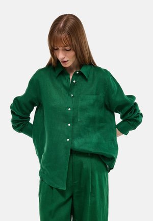 Femme aux cheveux auburn lisses portant une chemise boutonnée vert foncé ample et un pantalon assorti, regardant vers le bas, les mains derrière le dos.