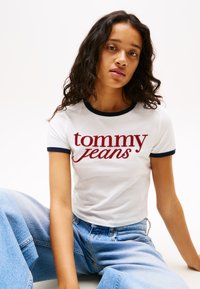 Fehér, rövid ujjú póló tengerészkék bordás nyakkivágással. A piros írásban "tommy jeans" logó található rajta. Világoskék, bőszárú farmernadrággal párosítva.