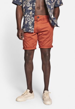 Mand iført rullede, brændt orange shorts, en marineblå blomster kortærmet skjorte, beige sneakers, står med den ene hånd i lommen.