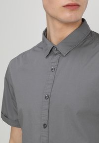 Grijs kortom mouw shirt met een gestructureerde kraag, gestikte details en drie zichtbare knopen aan de voorkant. Gladde textuur.