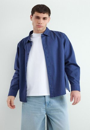 Młody mężczyzna stoi ubrany w niebieską rozpiętą koszulę na białej koszulce i jasnoniebieskich dżinsach na jednolitym tle.