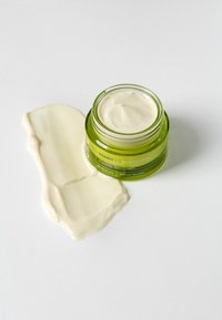 Grüner Glasbehälter mit einer weißen Creme darin, neben einem Klecks der Creme auf einer weißen Oberfläche. Text auf dem Behälter gibt Produktdetails an.