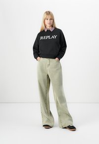 Zwarte sweatshirt met "REPLAY" in het wit gedrukt, gecombineerd met lichtgroene wijde jeans en zwarte schoenen. Heeft een geruite kraag eronder.