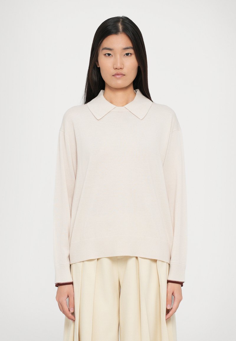 Maglione in maglia beige chiaro con un ampio colletto, spalle scese e polsini a coste. Abbinato a pantaloni plissettati in crema chiaro.