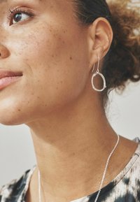 Pendiente de aro de plata con una superficie texturizada y un suave brillo, combinado con una delicada cadena de collar sobre un fondo neutral.