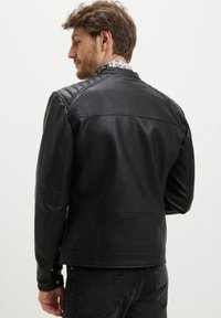 Giacca di pelle nera con dettagli cuciti sulle spalle; ha una texture liscia e una vestibilità sartoriale. Presenta un dettaglio con bottone a pressione sui polsini.