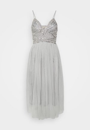 Srebrna sukienka bez rękawów z górą ozdobioną cekinami i głębokim dekoltem, przezroczysta, warstwowa spódnica z tiulu sięgająca poniżej kolan.