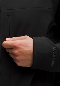 Juodas švarkas su užtrauktuku kišene ir Lululemon logotipu ant rankovės. Medžiaga yra lygi ir patvari, su prigludusia forma.