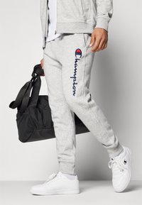 Grå joggebukser med blå "Champion"-logo, ribbestrikkede mansjetter, sammen med hvite joggesko. En svart treningsbag med doble stropper holdes i hånden.