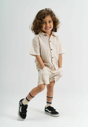 Glimlachend jong kind met krullend haar, gekleed in een beige gestreept shirt en shorts, met handen in de zakken, staand met één been gekruist, zwarte sneakers.