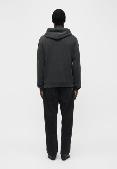 Άνδρας που στέκεται με την πλάτη, φοράει μαύρο φούτερ με κουκούλα και χαλαρά μαύρα τζιν μπροστά σε απλό λευκό φόντο.