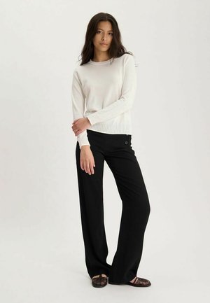 Femme debout, bras croisés, portant un pull blanc à manches longues, un pantalon noir à jambes larges et des chaussures tressées foncées.