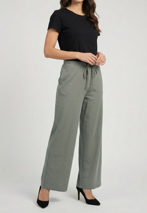 Femme portant un haut noir à manches courtes, un pantalon large gris clair avec une ceinture nouée à la taille, et des chaussures à talons hauts noires, debout les mains jointes.