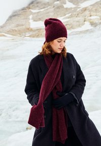 Burgundy knit beanie and scarf paired with a dark overcoat, featuring a textured finish and long, relaxed fit against a snowy backdrop.