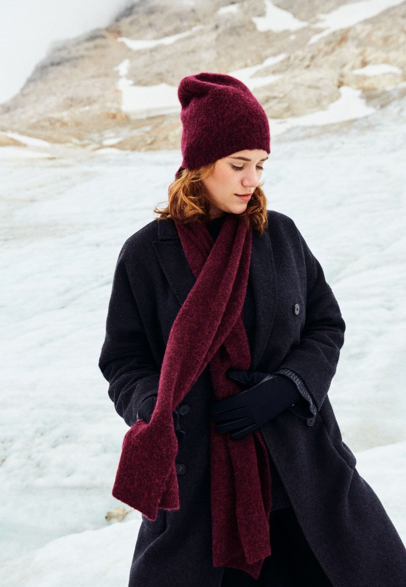 Burgundy knit beanie and scarf paired with a dark overcoat, featuring a textured finish and long, relaxed fit against a snowy backdrop.