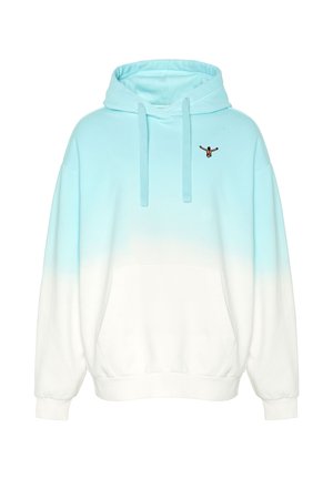 Mann trägt einen blau-weißen Kapuzenpullover mit Farbverlauf, dunkle Cargo-Shorts und weiße Sneakers, steht mit einer Hand in der Tasche und einer Hand am Kordelzug der Kapuze.