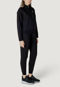 Tuta sportiva nera con giacca a zip e pantaloni aderenti; texture liscia, design fitted, abbinata a sneakers nere con suole bianche.