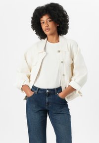 Lys kremfarget oversized denimjakke over en hvit skjorte, sammen med mørkeblå jeans. Jakken har tre knapper foran og klaftlommer.