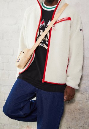 Homme portant une veste polaire blanche avec une bordure rouge, un pull graphique noir, un jean bleu et un sac beige porté en bandoulière, debout contre un mur en briques blanches.