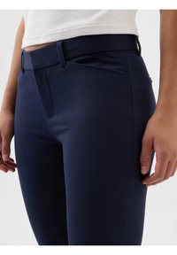 Gros plan d'une personne portant un pantalon bleu marine ajusté et un haut blanc, les mains détendues le long du corps sur un fond uni.
