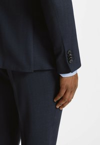 Main reposant à côté d'un pantalon de costume bleu foncé et d'une manche de veste avec quatre boutons noirs sur un poignet de chemise bleu clair.