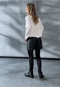 Modella che indossa un maglione bianco oversize, pantaloni shorts in pelle nera, collant neri e stivali alla caviglia con fibbie. Sfondo in cemento liscio.