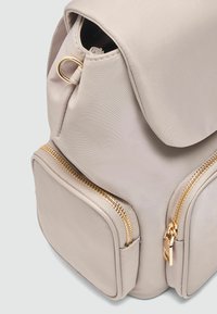 Sac à dos beige clair en tissu lisse avec des zips dorés. Il possède des bords arrondis et deux poches extérieures zippées.