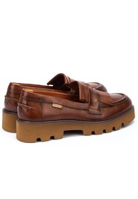 Mocasines de cuero marrón con diseño de costura, flecos decorativos y una suela beige gruesa. Presentan una etiqueta de logo en el lateral.
