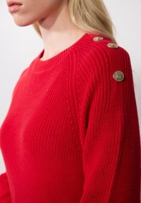 Pull en tricot rouge avec une texture côtelée, encolure ronde et boutons dorés à l'épaule. Le design comprend des manches longues et une coupe ajustée.
