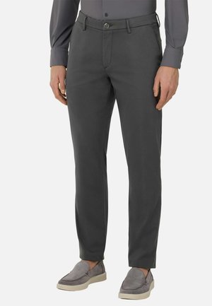 Donkergrijze op maat gemaakte broek met een platte voorkant, riemlussen en een ritssluiting. Gemaakt van een gladde stof; gedragen met een overhemd met lange mouwen en grijze schoenen.