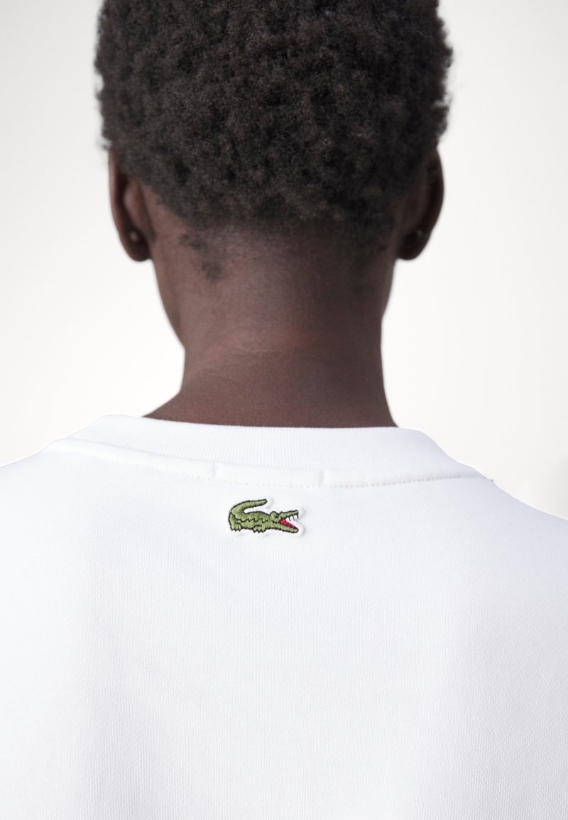 Polo blanc avec une finition texturée, présentant une petite broderie de crocodile vert au niveau de la nuque à l'arrière.